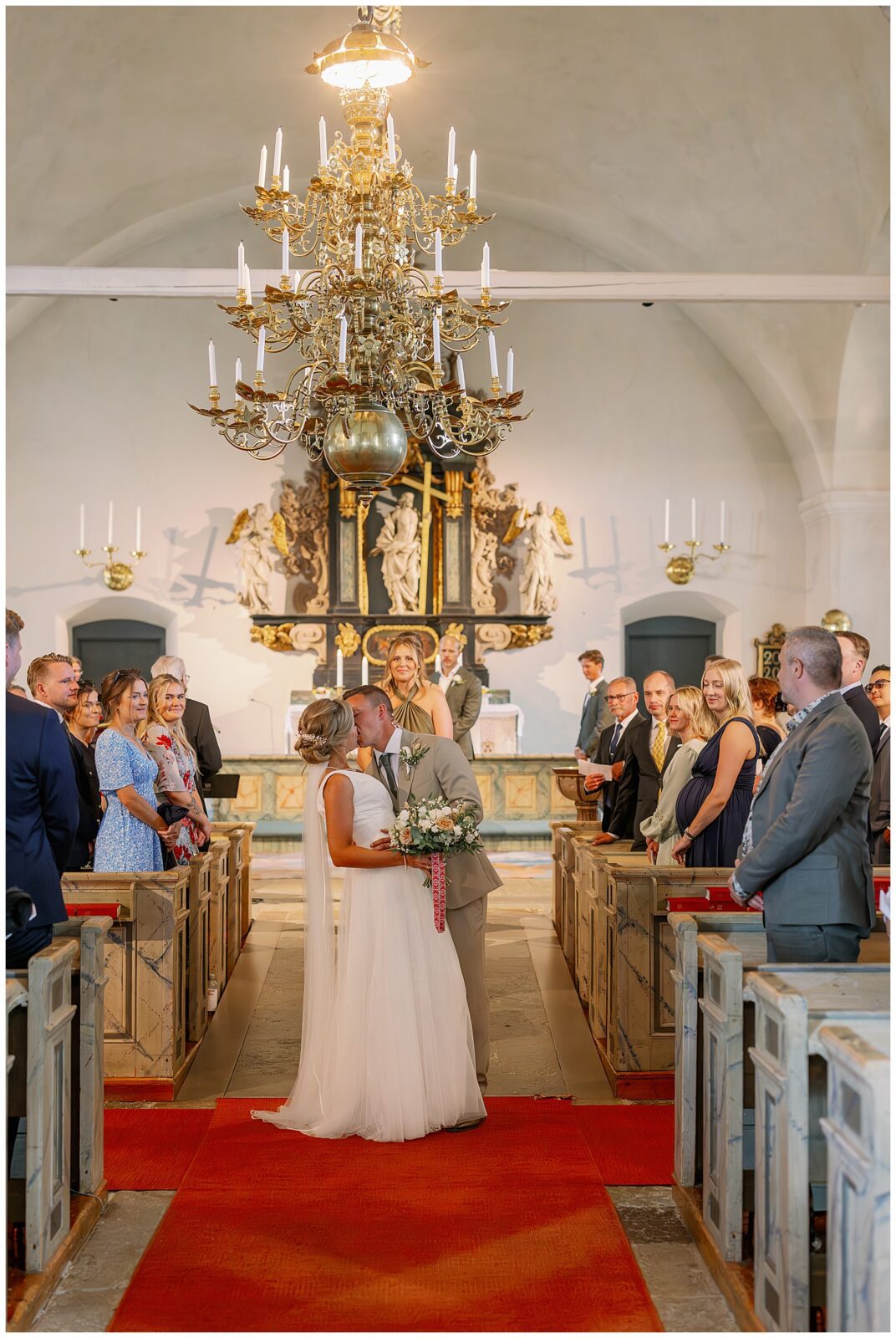 Brud och brudgum kysser varandra i mittgången i Rättviks kyrka, omgivna av gäster | Bride and groom kissing in the aisle of Rättvik Church, surrounded by wedding guests