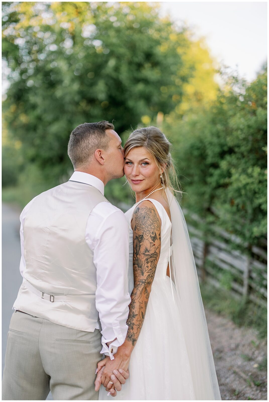 Närbild på brudpar som håller varandra i handen på en lugn väg i Tällberg, med brudens slöja och tatuerade arm i fokus, fångat i varmt golden hour-ljus vid Green Hotel. Bröllopsfotograf Dalarna med dokumentär känsla. Close-up portrait of a bride and groom holding hands on a quiet road in Tällberg, with the bride’s veil and tattooed arm in focus, captured in warm golden hour light at Green Hotel. Documentary wedding photography in Dalarna.